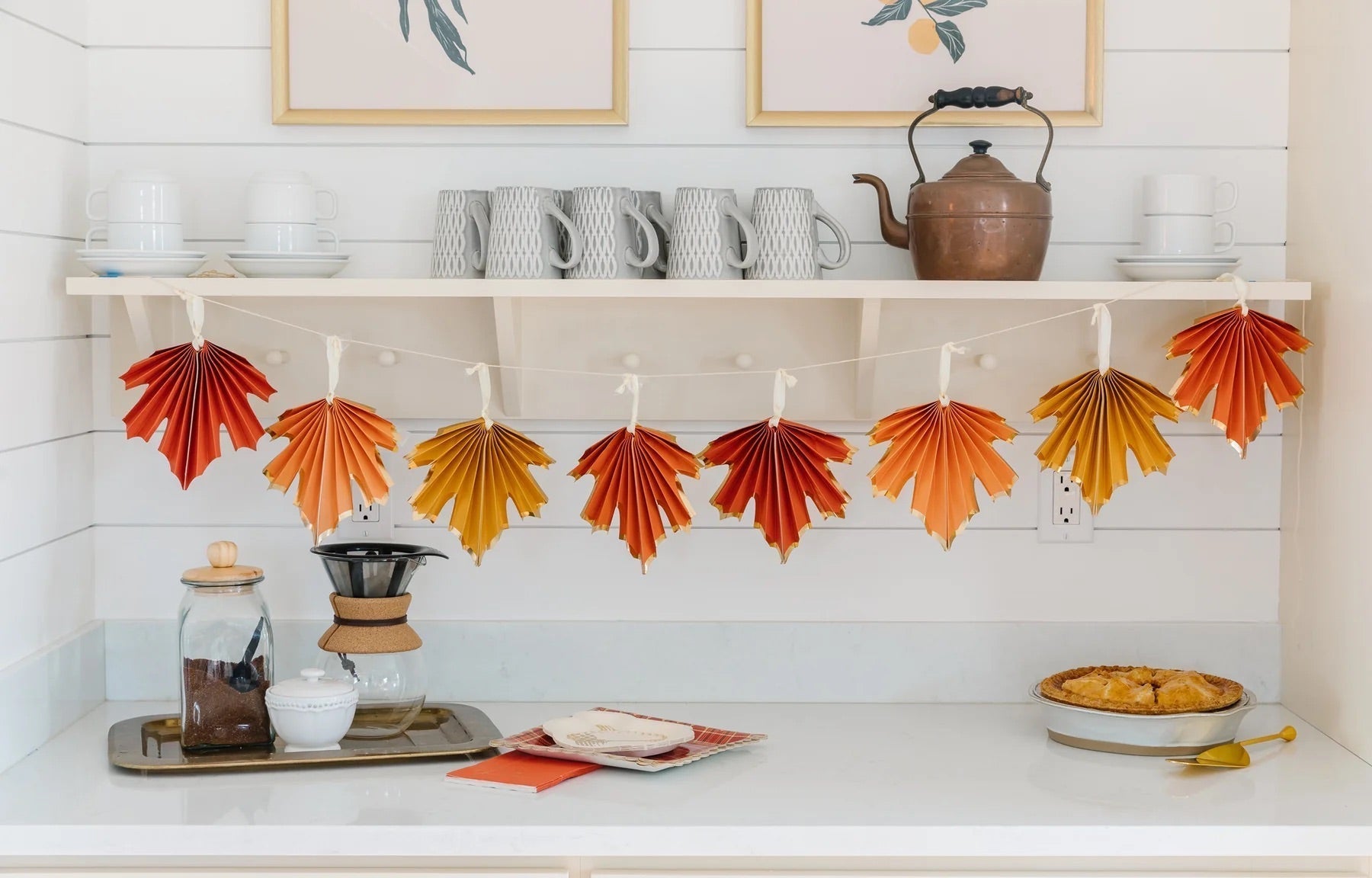 A kitchen counter decorated with maple leaf paper party banner from Momopartys. Underneath are some coffee brewing set and homemade baked pie, a very autumn fall home decor idea for the harvest season.