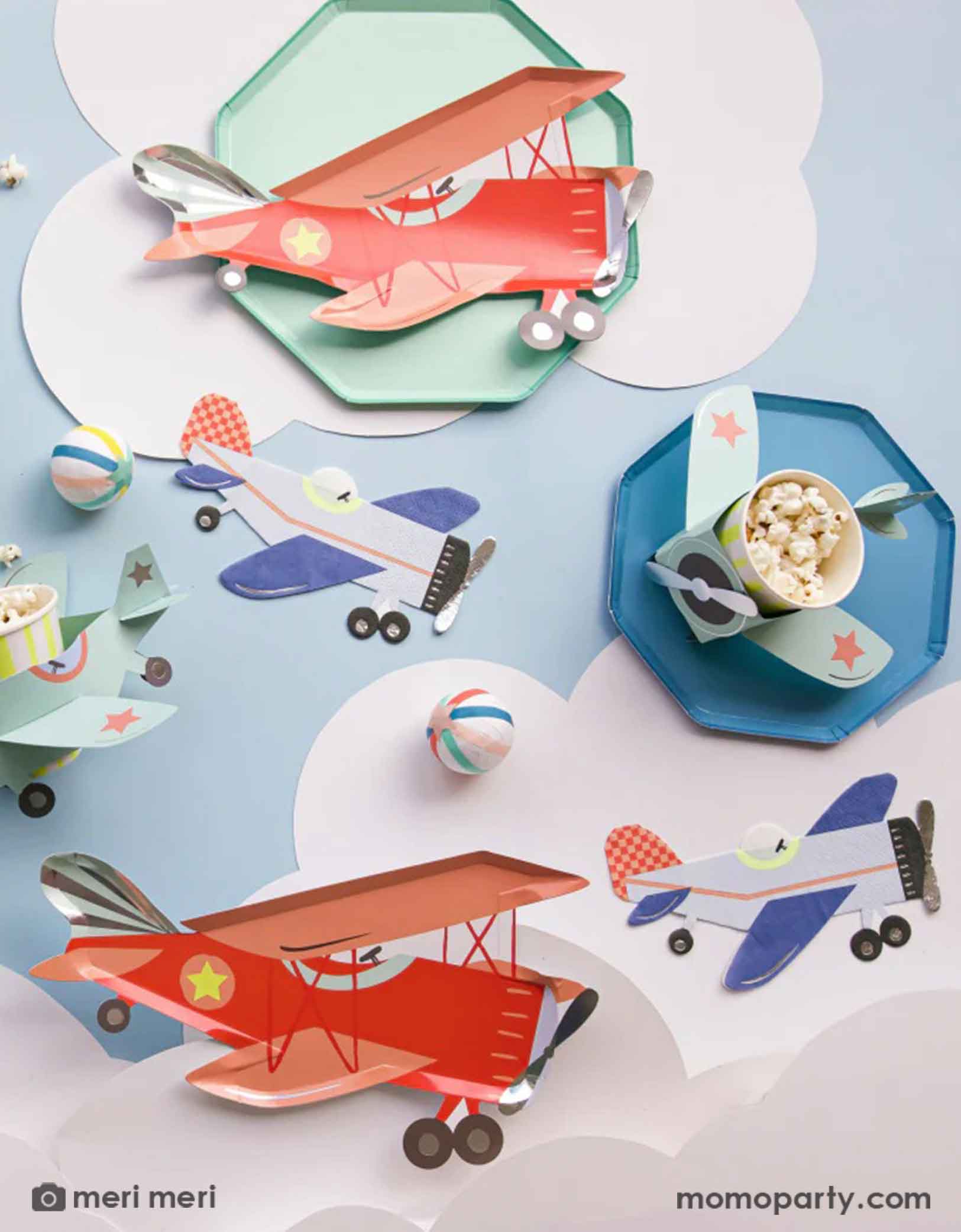 A table of fun and adorable airplane themed party tableware from Momopartys by Meri Meri, including red retro vintage plane shaped paper plates, blue vintage plan shaped napkins and airplane 3D paper party cups, along with cloud paper cutouts, this look is a great inspiration for kid's airplane themed party decorations.