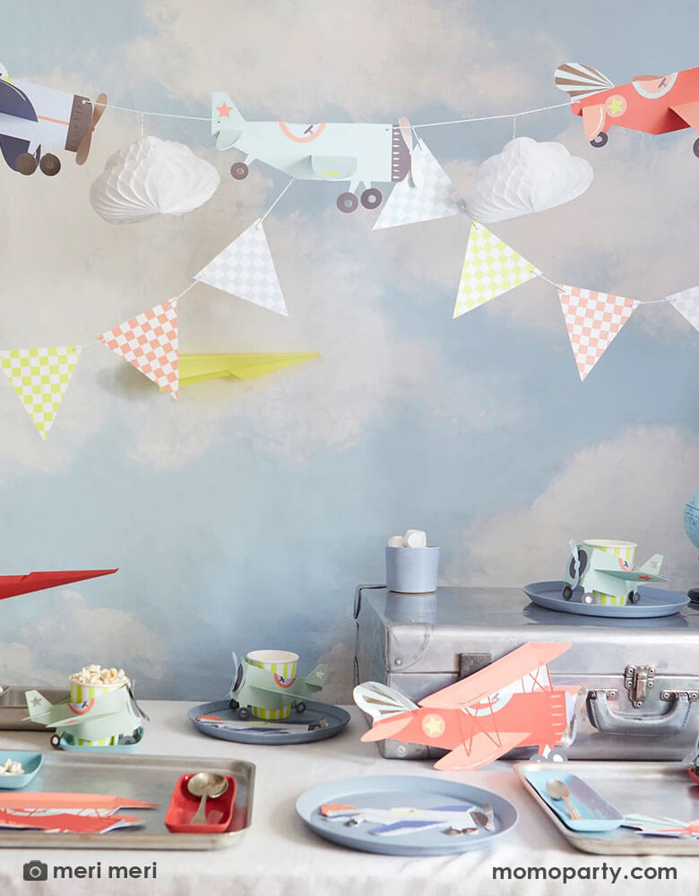 A airplane themed birthday party featuring Momopartys's plane themed decorations and party supplies by Meri Meri including red vintage airplane shaped plates, blue airplane shaped napkins and paper cups filled with drinks and treats. Above the table is a 3D honeycomb paper crafted plane garland against a blue sky with clouds wallpaper which sets a perfect scene for this airplane / aviator themed birthday party.