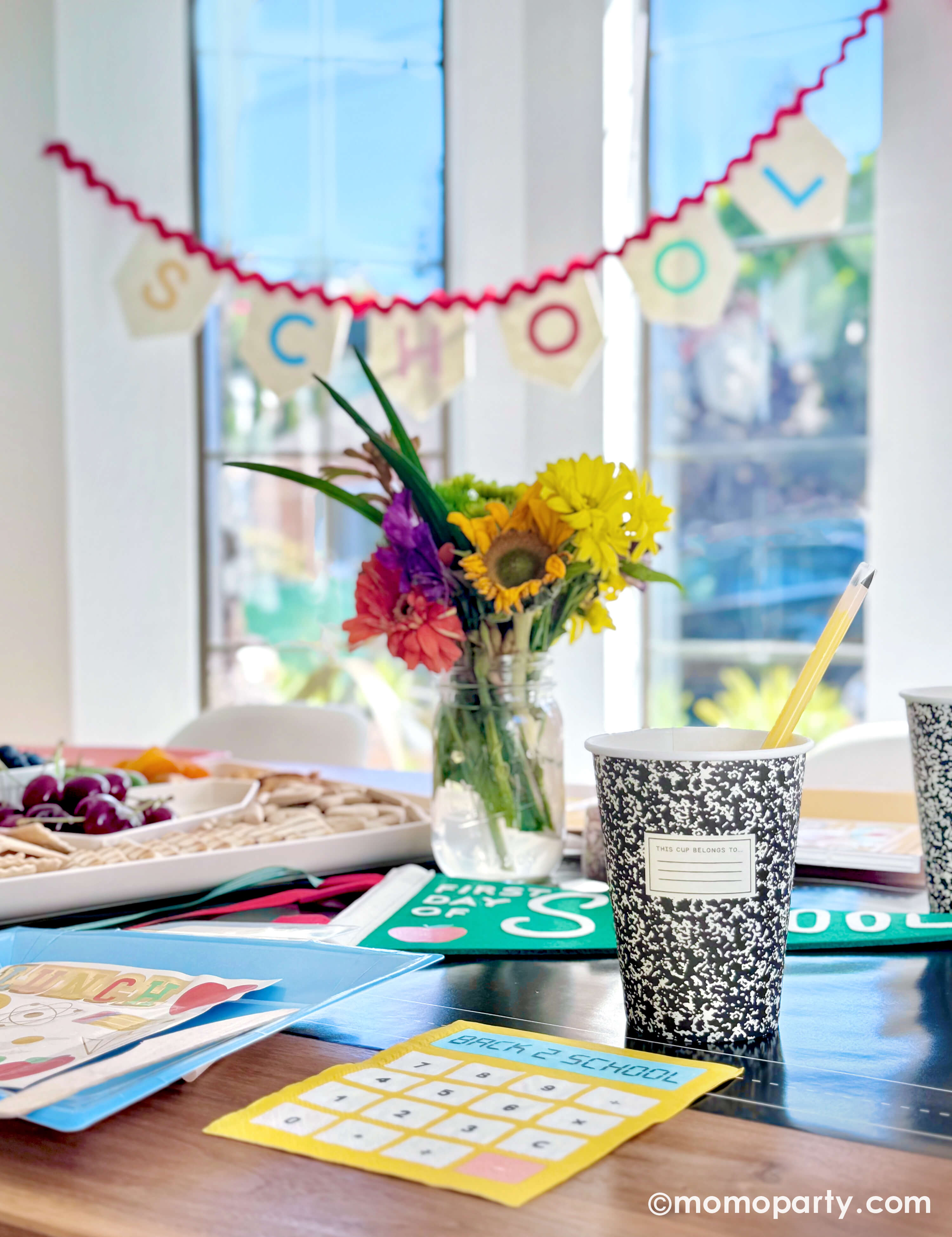 A kids' back to school party table setting featuring various school themed party decors and tableware from Momopartys including notebook party cups with pencil reusable straws, calculator shaped dinner napkins, lunchbox shaped plates and apple first day of school party pennant, with a summer flower arrangement and a kid's snack board, along with "School Day" party garland, this simple yet festive set up is perfect to celebrate your child's first day of school!