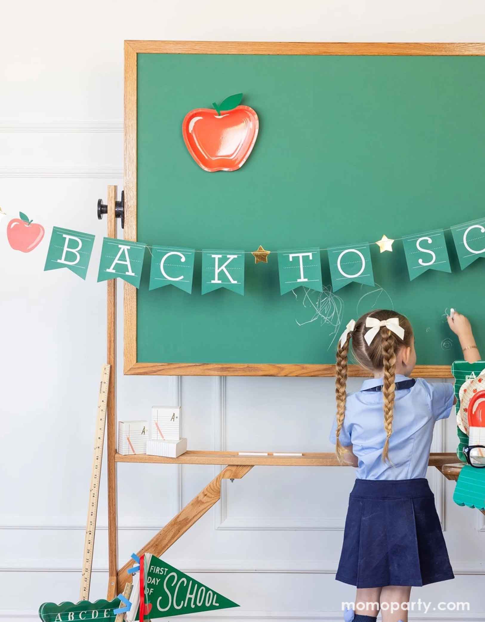 A school-aged girl in her blue uniform writing something on a chalkboard in a classroom set up decorated with a bunch of school themed party supplies from Momopartys including 8.5" apple shaped plates, 10" apple pattern plates, alphabet dinner plates and matching napkins. The chalkboard is adorned with the back to school party banner hung across, with an apple shaped plate on it - an adorable and cute set up to celebrate kid's back to school season!