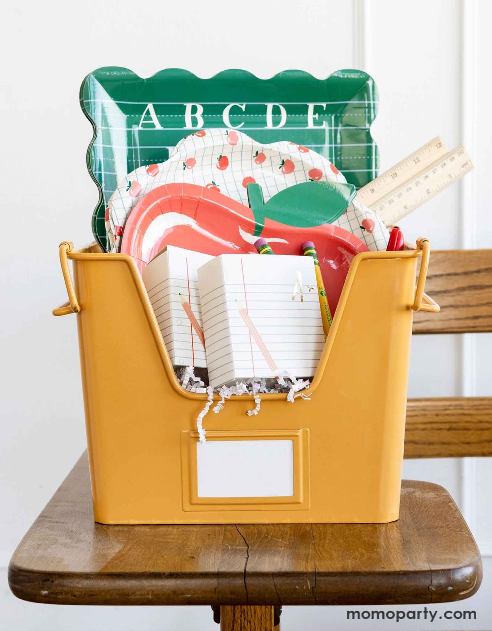 A vintage inspired yellow file bin filled with various school themed party supplies from Momopartys's including two ruled paper treat boxes, an apple shaped paper plate, an apple patterned scallop-edged plate and an ABC alphabet green dinner plate. The bin is placed on a classic classroom chair - a perfect inspiration for celebrating kid's back to school season or first day of school occasion!