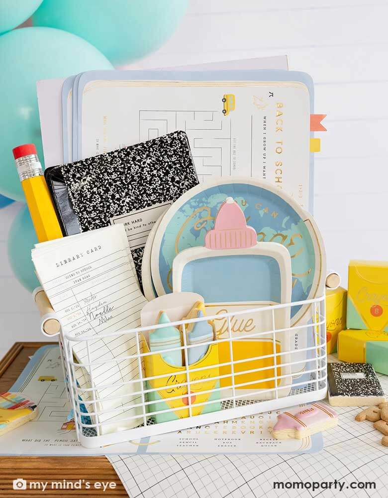 A back to school occasions bin filled with school themed party supplies from Momopartys including back to school planner placemats, globe shaped plates, glue bottle shaped plates, library card shaped napkins, a crayon box shaped treat box filled with crayon shaped sugar cookies and a giant pencil. On the table there's a notebook grid inspired table runner. With the party balloons in the back, it's a great inspo for a festive kid's first day of school celebration party!