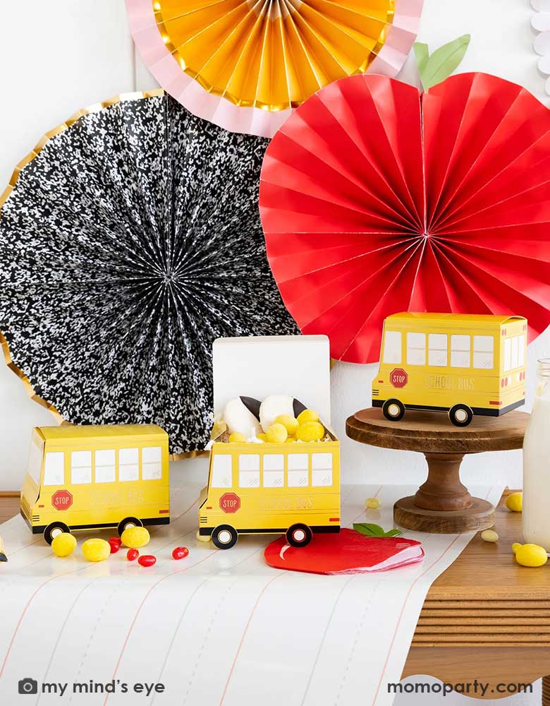 A festive back to school party table features Momopartys's school themed party supplies including school bus shaped treat boxes, apple shaped napkins, writing paper table runner by My Mind's Eye. On the table are some red jelly beans, yellow candies and milk bottles in coordinating colors. In the back there's the back to school party fan hung on the wall, making this a perfect inspiration for kid's back to school party or a first day of school celebration breakfast.