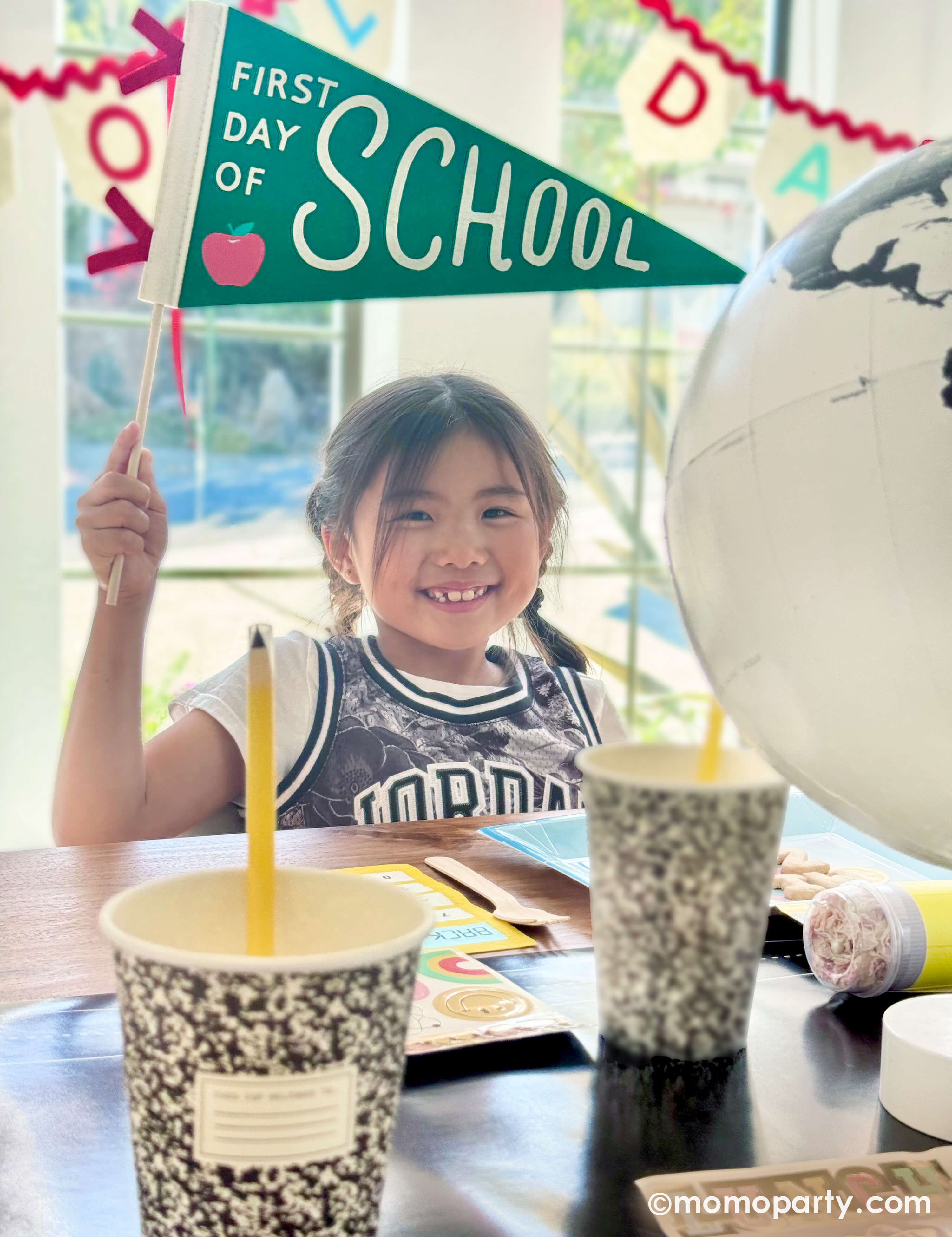 A school aged girl at a back to school celebration holding Momopartys's first day of school apple felt party banner sitting at a table decorated with various of school themed decorations and party tablewares from Momopartys including the chalkboard table runner, notebook party cups, pencil confetti poppers, lunchbox shaped party plates and school day felt party garland. A perfect inspo for your child's first day of school celebration!
