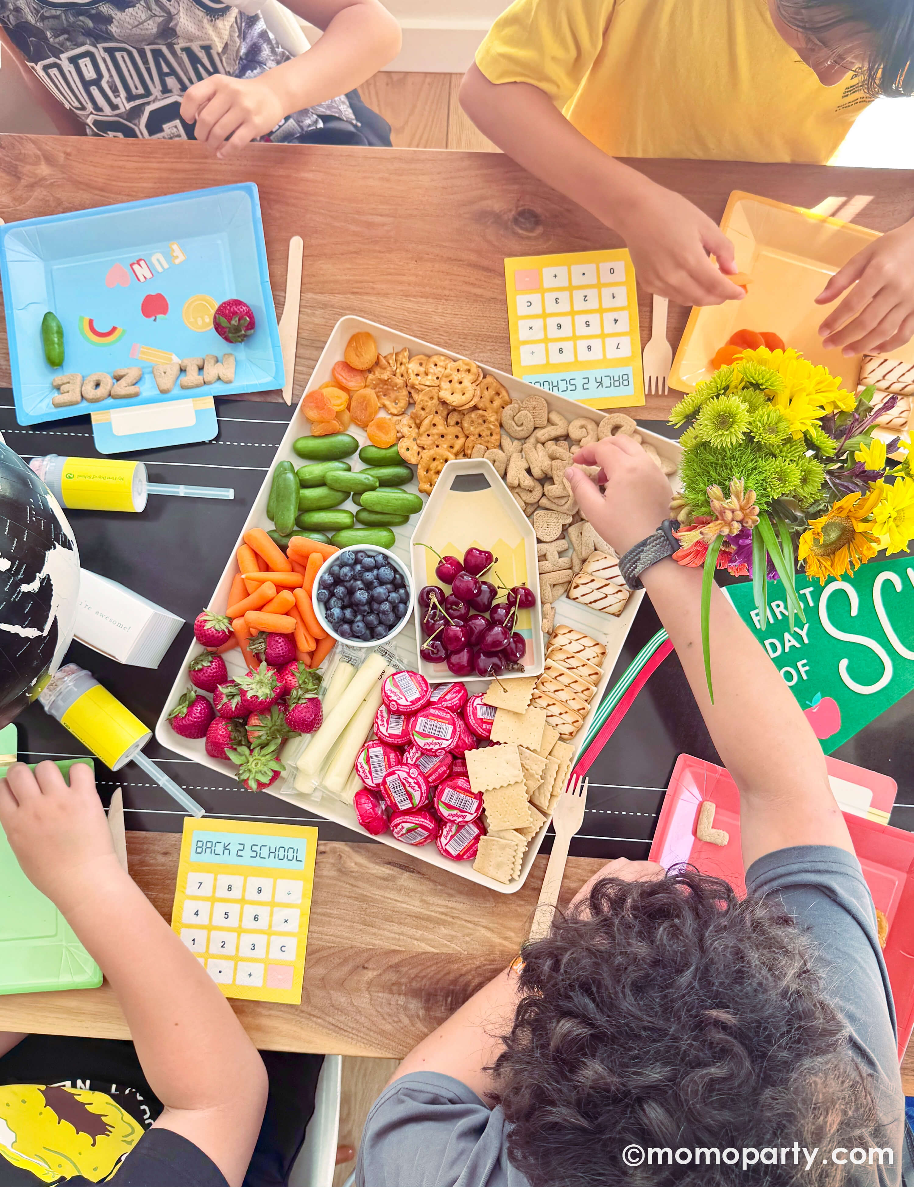 A few kids sitting around a festive back to school party table set up featuring various school themed party decors and tableware from Momopartys including the chalkboard table runner, lunchbox shaped party plates, calculator shaped napkins, notebook party cups. In the middle of the table is the paper and pencil bamboo tray set which is filled with healthy snacks including fruits, veggies, cheese and cookies for kids to grab. What a fun idea for kid's back to school playdate to celebrate the new school year!