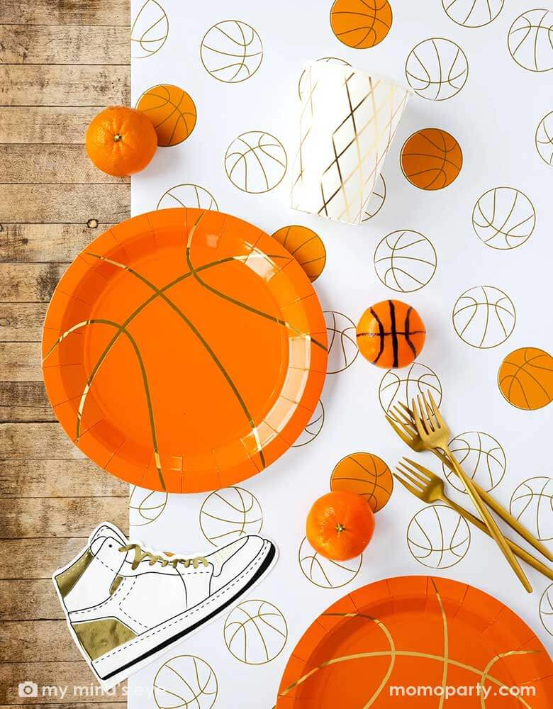 A basketball themed party table features Momopartys's basketball tableware by My Mind's Eye including basketball shaped plates, hightop sneakers shaped napkins, basketball net gold foil party cups and paper basketball table runner, with oranges as snacks on the table - a perfect table setting inspo for kid's basketball themed birthday parties or a fun NBA viewing party!