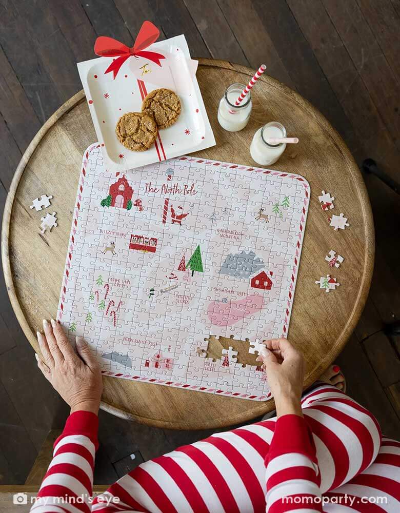 A lady in her red and white striped pajama playing a Christmas themed puzzle next to Momopartys's Believe Santa Present with a bow paper plates with cookies in them, next to it are two bottles with milk with red striped straws, creating a Holiday cozy vibe at home.