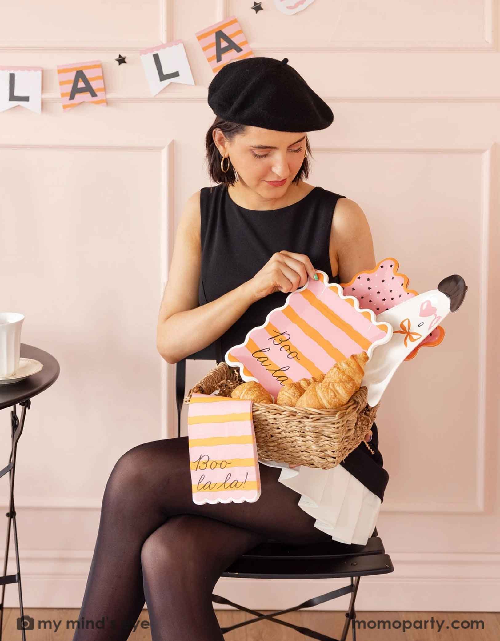 A lady in a black dress and beret holding a basket with Momopartys's Boo La La french inspired Boo La La party tableware collection featuring the ghost-shaped plate, the orange and pink striped plate and napkins the at a Halloween-themed party decorated with french-inspired striped Boo La La party banner on the wall.