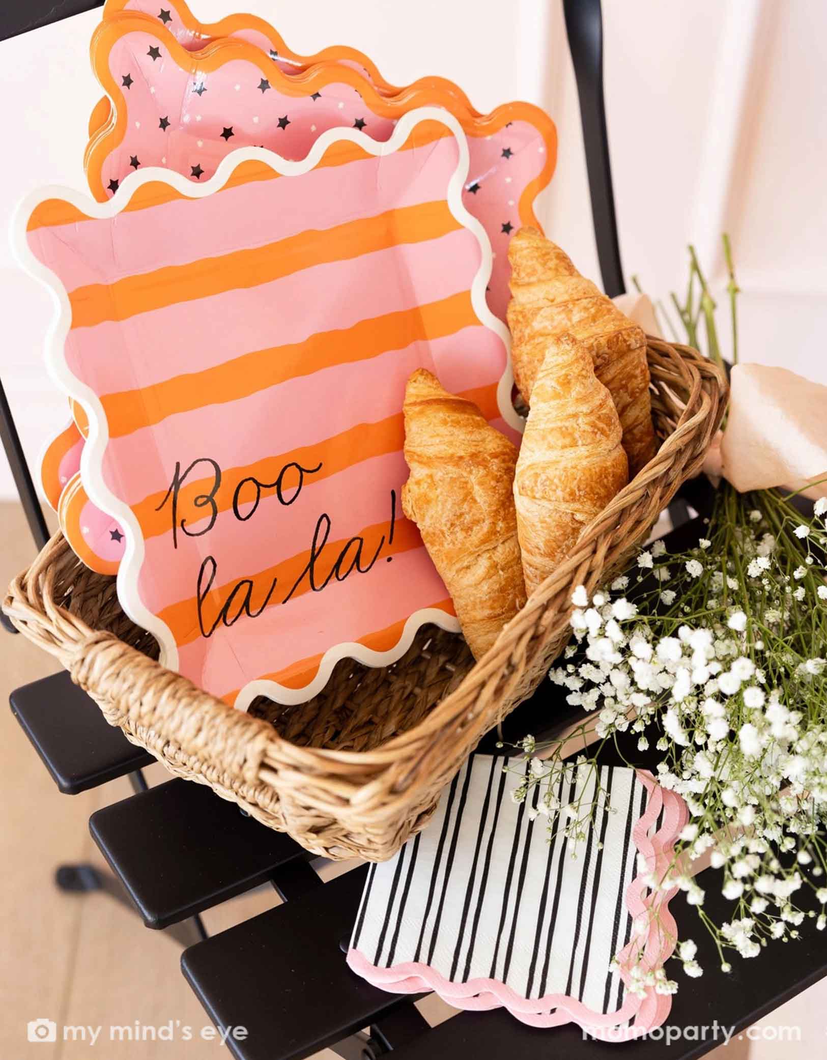 A french inspired basket filled with Momopartys's Boo La La party tableware collection including the orange and pink striped dinner plates, the pink and black star plates, black and pink striped napkins and with pastries and flowers around it.