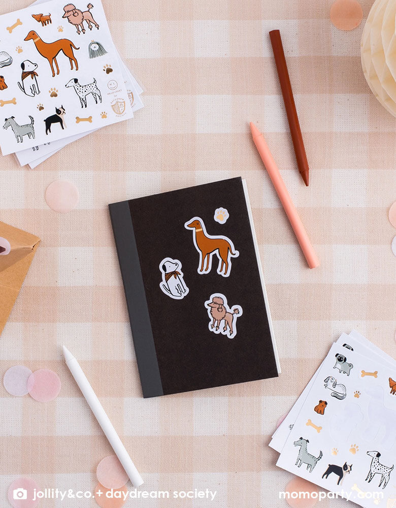A soft peach checkered table with a small black notebook with Momopartys's Bow Wow dog stickers on it. Around the notebook there are nude/natural colored pencils, giving some inspirations for kid's puppy/dog themed birthday party goodie bag filler ideas.