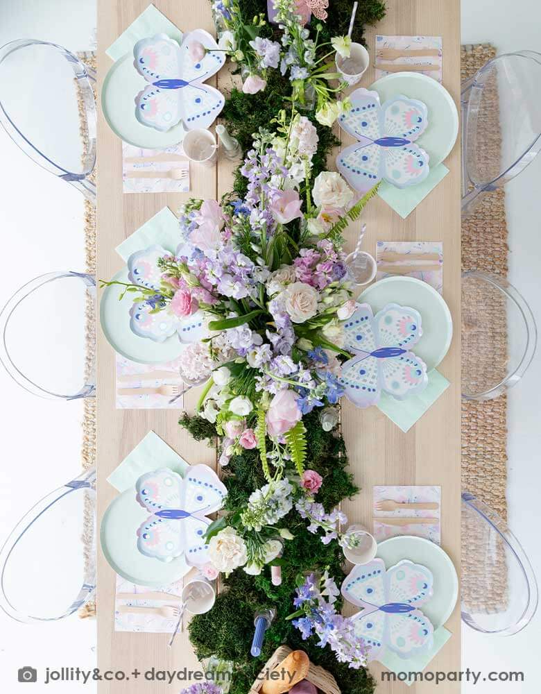 An overhead shot of a beautiful butterfly themed party table featuring Momopartys's Flutter butterfly shaped plates paired with pastel mint dinner plates and napkins, next to it is butterfly themed party cups and napkins. In the middle there's moss grass textured table runner with pretty spring flower arrangement. On the wall there are butterfly shaped paper decorations attached on the wall, making this a perfect inspo for kid's butterfly fairy themed birthday or a gorgeous spring Easter celebration.