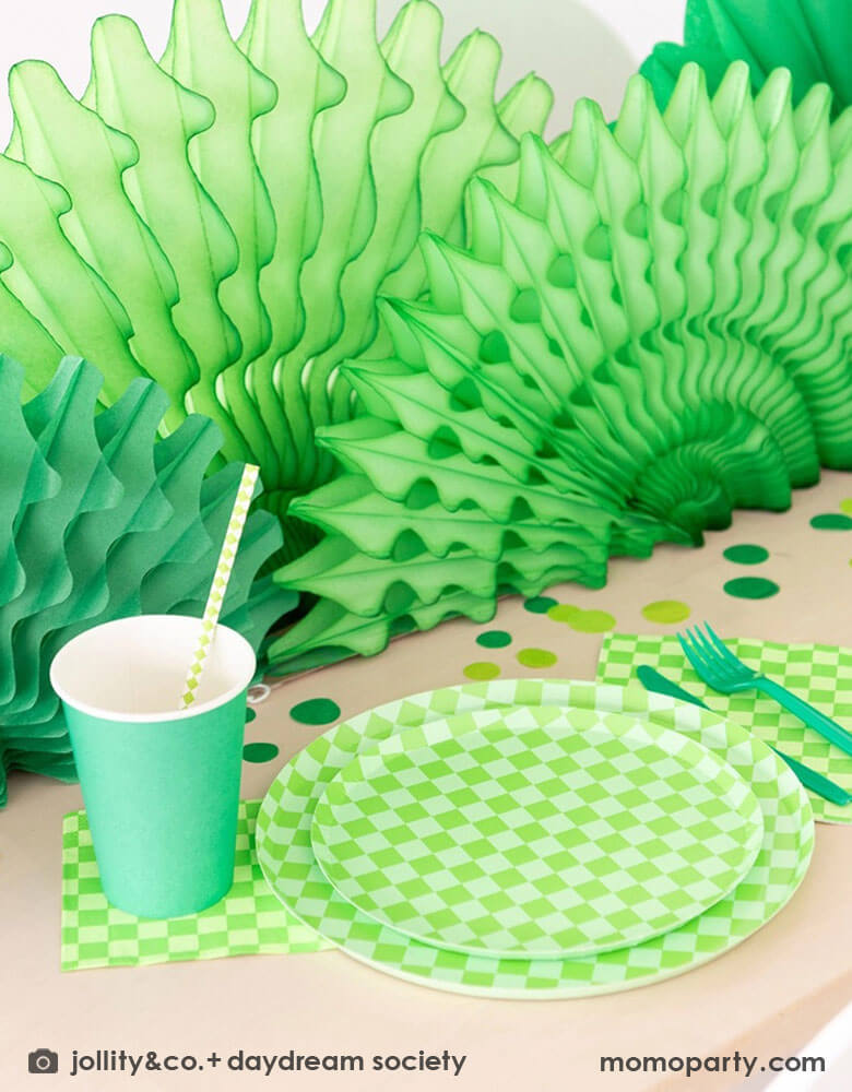A green themed party table features Momopartys's Check it! Lime Green Checkered Tableware Collection by Jollity & Co. including 10" dinner plates, 8" dessert plates, 6.5" large napkins, green utensils and solid green party cups, with green paper fans as the centerpiece and large round green confetti scattered on the table.