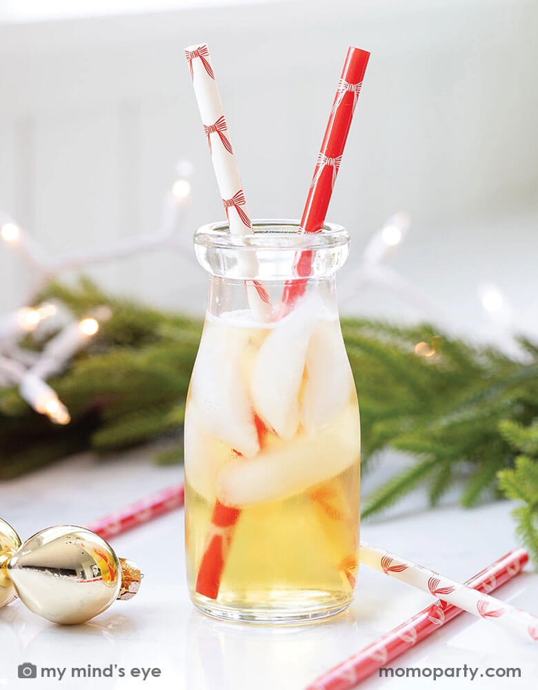 A close up of a glass filled with apple juie with Momopartys's red and white Christmas Bows reusable straws by My Mind's Eye. Around the table there's a Christmas wreath and some Christmas tree ornaments in gold. Making this a festive vibe for the Holiday season.