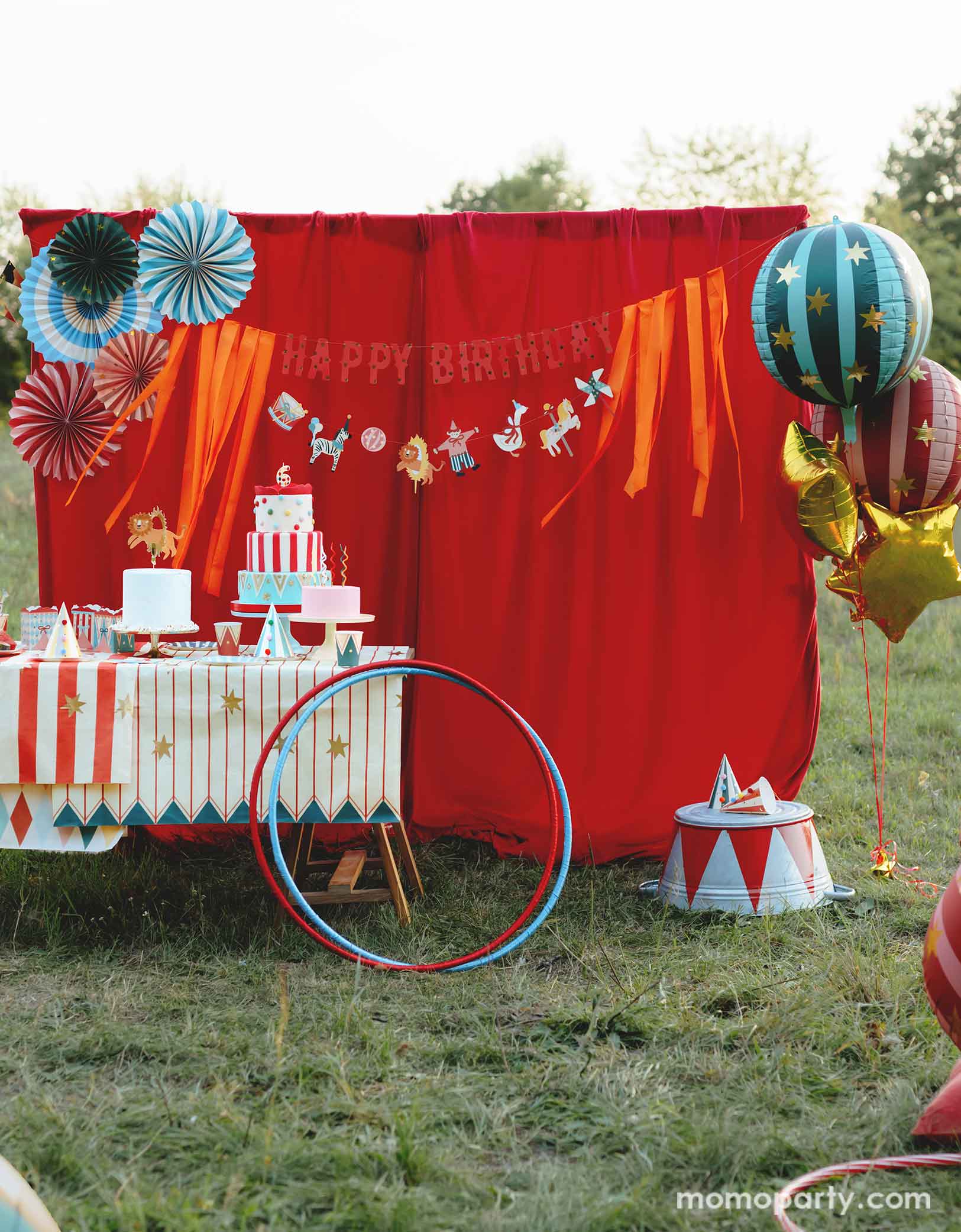 A vintage circus themed party set up in the park featuring various carnival circus themed party decorations from Momopartys including the red and green striped globe foil balloons along with gold star foil balloons, a red stage curtain decorated with Happy Birthday circus banner and colorful paper fans and a table filled with party tableware and multiple birthday cakes. Making this a whimsical and playful setting for a fun kid's circus birthday party.