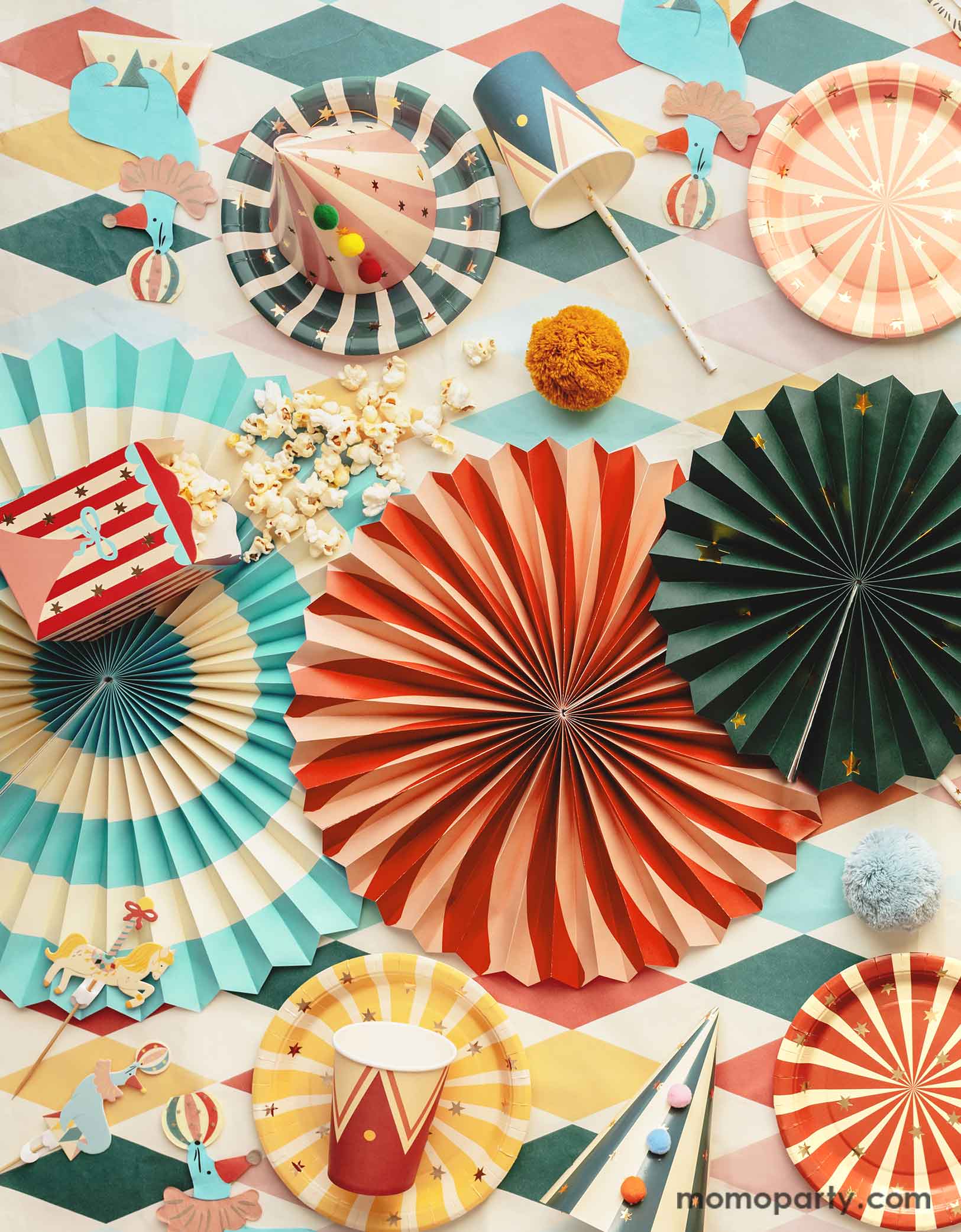 A carnival/circus themed part table featuring Momopartys's circus themed party supplies including colorful striped paper plates in 6 different colors, sea lion playing a ball shaped napkins, matching party cups, treat boxes, striped party hats with pom poms, circus themed cake toppers, and festive paper fans, with gold foil straws and popcorns on a circus themed striped table cover - making this great table setting idea for a fun and festive kid's carnival themed birthday celebration!