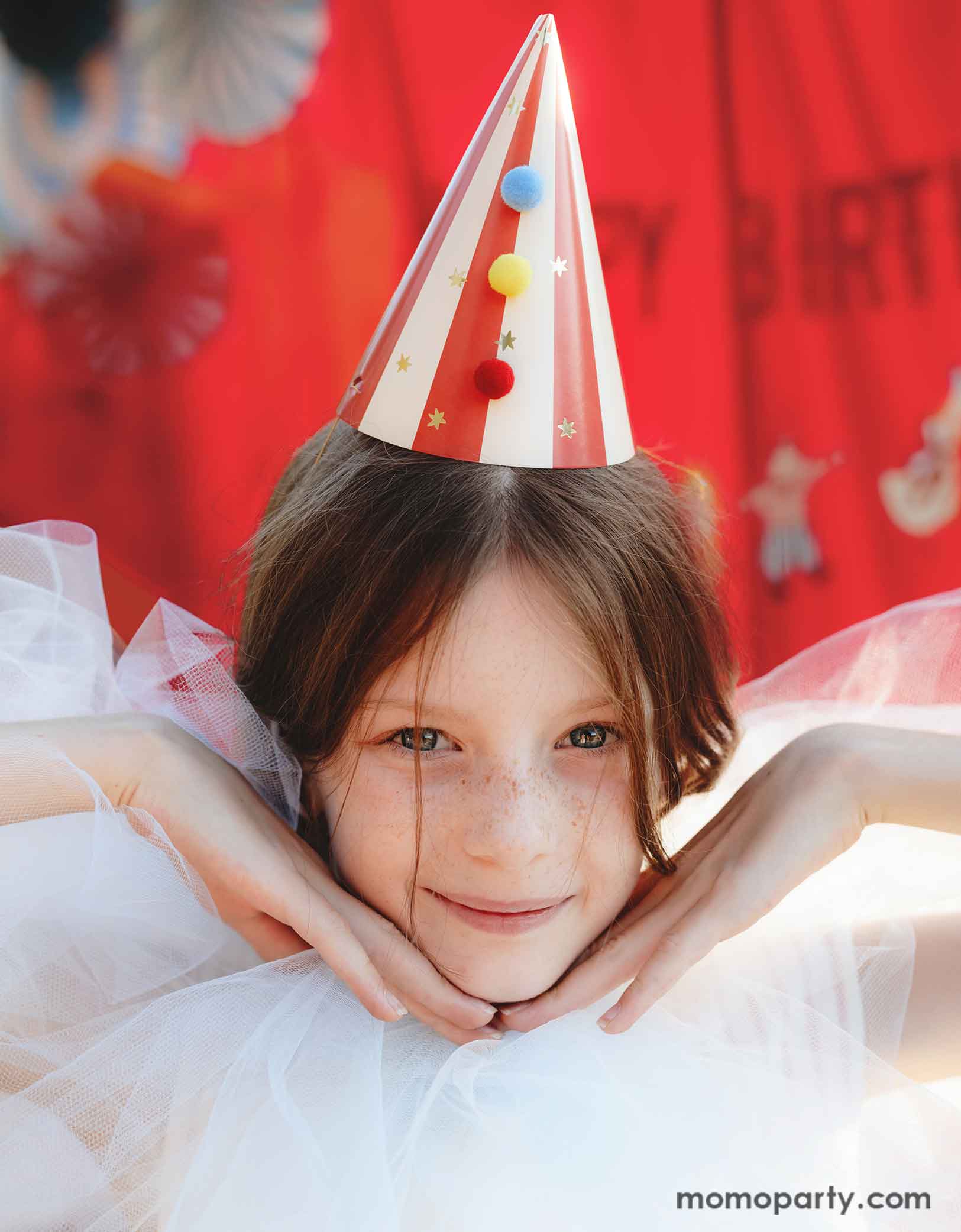 A girl dressed in dreamy white tulle dress wearing Momopartys's red striped Birthday Party Hat with Pompoms, in a festive carnival themed event. In her back is a red stage curtain decorated with circus themed birthday banner and colorful party fans.