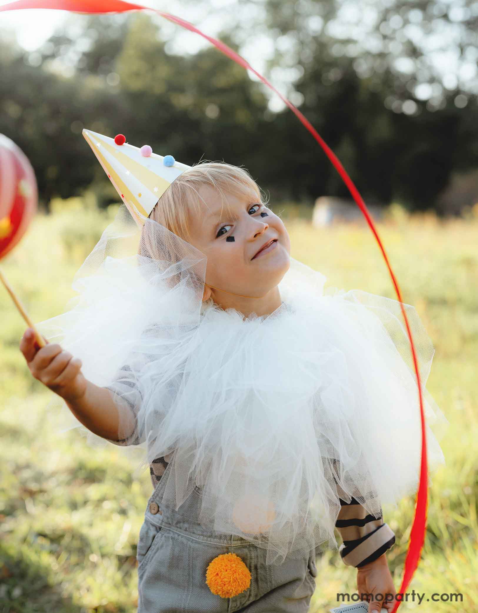 A little boy dressed in dreamy white tulle cover with vintage circus themed outfits wearing Momopartys's yellow striped Birthday Party Hat with Pompoms, in a festive carnival themed event. He's waving a red party streamer in the field and having fun.