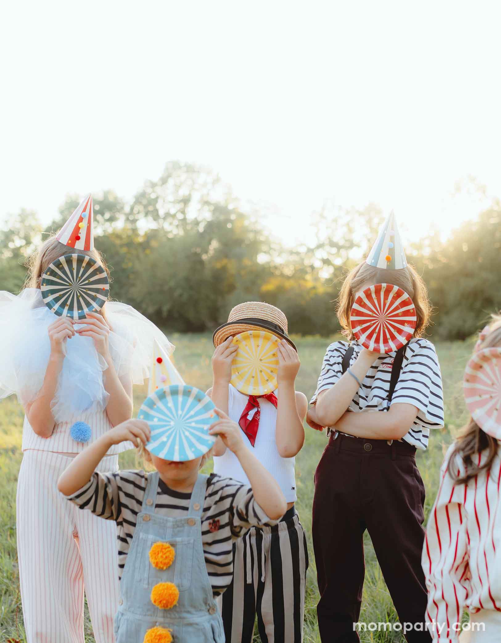 A bunch of kids dressed in the vintage circus outfits holding Momopartys's 7" Circus Striped Plates covering their faces. This set of 6 plates features vibrant stripes in 6 different colors and a charming gold star design, adding a touch of carnival magic to any meal. They're perfect additions to your circus or carnival themed party table!