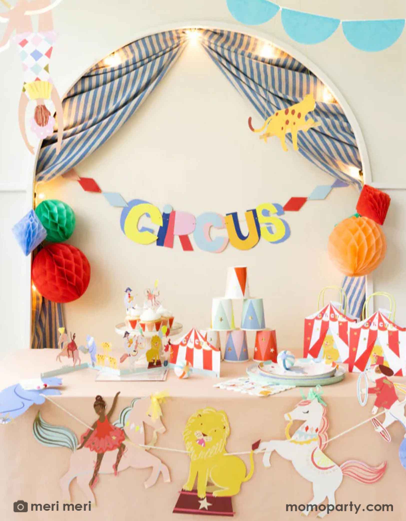 A festive circus themed party set up featuring Momopartys's circus themed party supplies including a table filled with circus themed party tablewares including plates, napkins, party cups, party bags and ferry's wheel cupcake stand. There's the circus party garland featuring circus animals with honeycomb decorations hung above the party table.