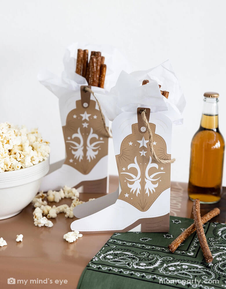 A cowboy themed party table filled with treats and snacks and Momopartys's cowboy boot shaped treat boxes filled with pretzel sticks. On the table there's a soda bottle and a green bandana as decoration, perfect for a Western rodeo themed celebration, be a First Rodeo boy's first birthday party or a fun cowboy bash!