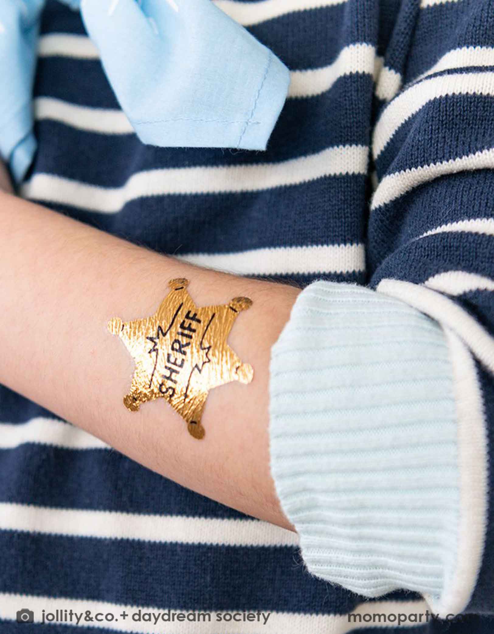 A boy wearing navy striped shirt with a light blue bandana. On his arm he has Momopartys's sheriff badge temporary tattoo on it. It's a great party favor for kid's Western or cowboy themed birthday party.