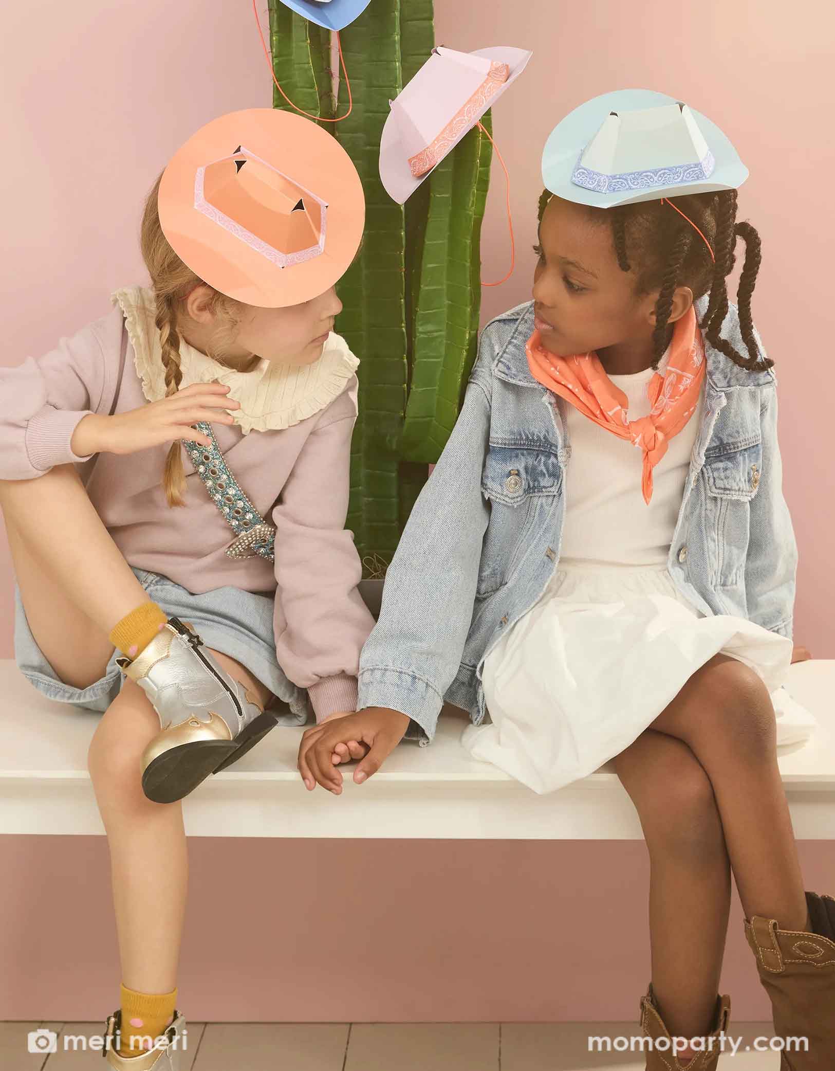 Two school aged girls dressed in Western inspired outfits including bandanas and cowboy boots, sitting on a white bench in front of cactus plant. Each of them wear the Meri Meri's 6.3" mini Western party hat, one in light blue, one in orange. There are also a couple more hats hung on the cactus plant.