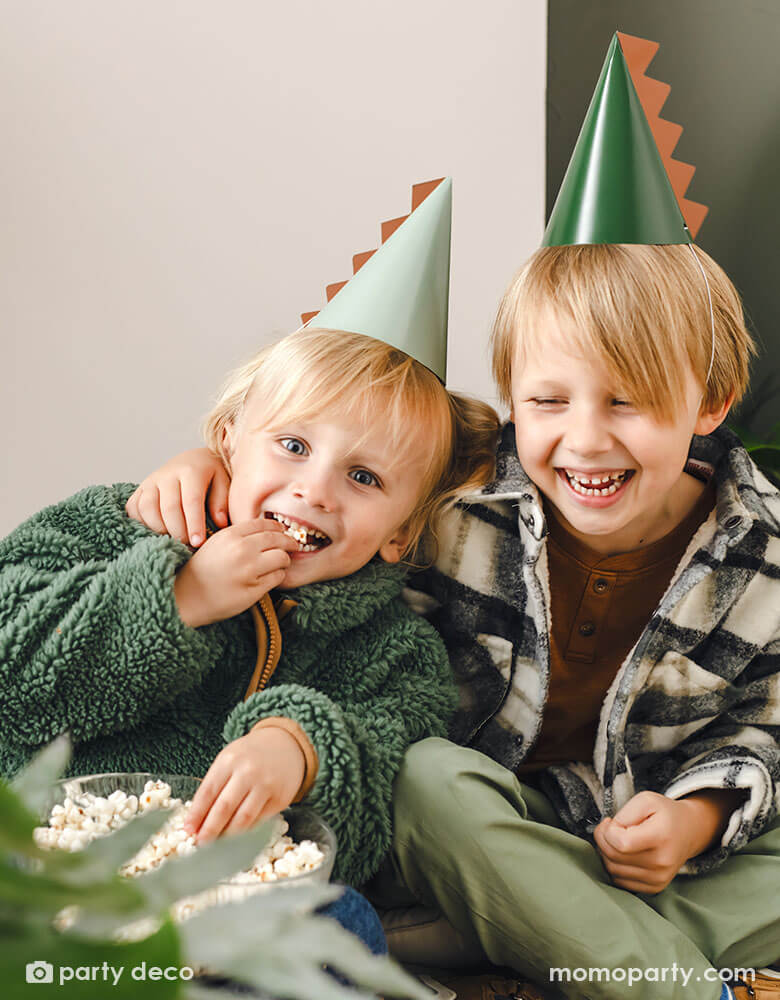 Two school aged boys in green themed outfit wear Momopartys's 5.5" Dinosaur Party Hats with a dinosaur spike on them by Party Deco. They're enjoying the popcorn from the party, laughing and having a good time.