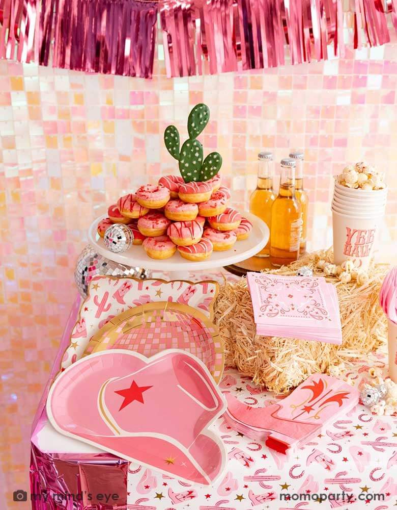 A cowgirl rodeo Western themed party table features pink cowgirl themed party supplies from Momopartys including pink cowgirl boot shaped napkins, cowgirl hat shaped plates, pink disco ball shaped plates, pink bandana small napkins, yeehaw party cups and cowgirl pattern paper plates and table runner by My Mind's Eye. In the back there's an iridescent backdrop adorned with pink foil fringe banner, along with pink donut tower cake topped with a cactus, making this a great inspo for a disco cowgirl party.