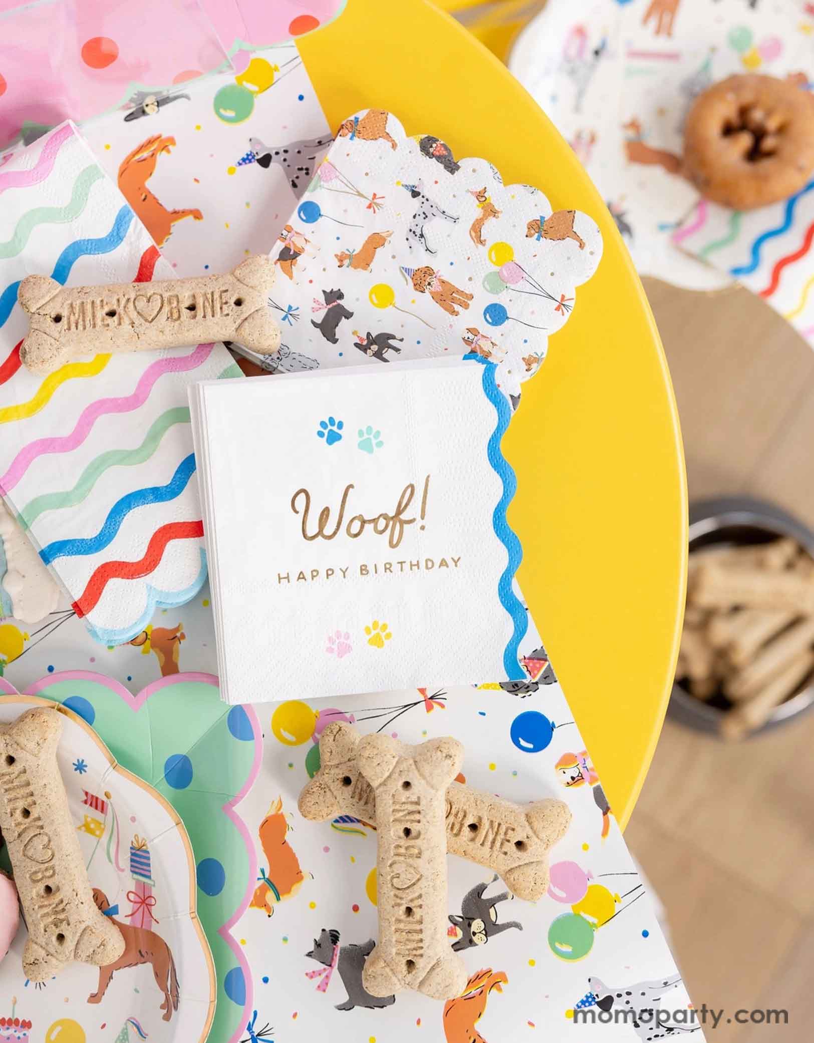 A festive and cheerful dog themed party table featuring Momopartys's dog party tableware collection including dog paper plates, woof! Happy Birthday napkins and table runner featuring adorable illustrations of different breeds of dogs with party hats, balloons and birthday cakes. With mint polka dot square plates and rainbow ric rac guest napkins, along with dog bone shaped cookies, this table is ready for a fun and playful birthday celebration for a pet lover!