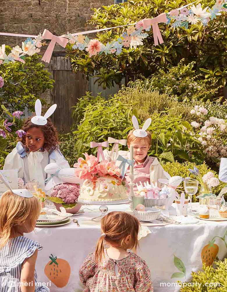 An adorable Easter party in the garden for kids featuring a long party table filled with Easter themed party supplies including bunny party cups, napkins, plates and party crackers. In the middle of the table, there's a large cake adorned with floral toppers and Momopartys's 3D bow cake toppers in pastel colors. Above the table hung the flowers and bows party garland, creating a wow factor to this gorgeous Easter celebration set up.