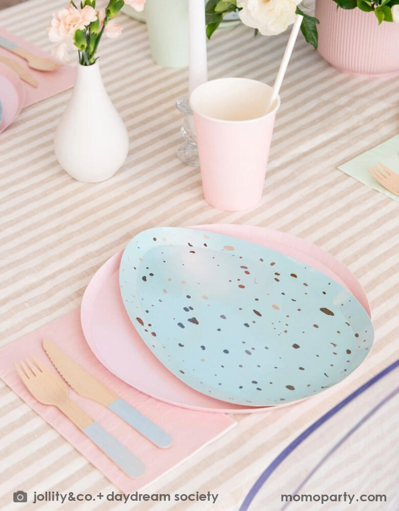 An closeup shot of Easter party table set up with Momopartys's Rockin' Robin Dinner Plates layered with pastel pink round plate, color matching paper cups, napkins, and pastel blue dipped wooden utensils, with vases with flowers and candles on a light beige striped tablecloth for a modern chic Easter table setting.