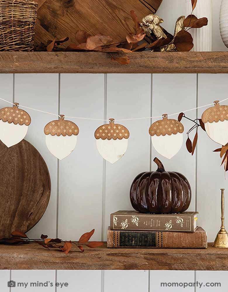 A fall inspired kitchen shelf decorated with Momopartys's 9ft wood acorn banner by My Mind's Eye. On the shelf, there are some fall colored table decorations including wooden cutting boards, books in brown covers, gold metal squirrel and broom decoration, some baskets and fall leaves spread around - a perfect inspo for fall home decors in this harvest season.