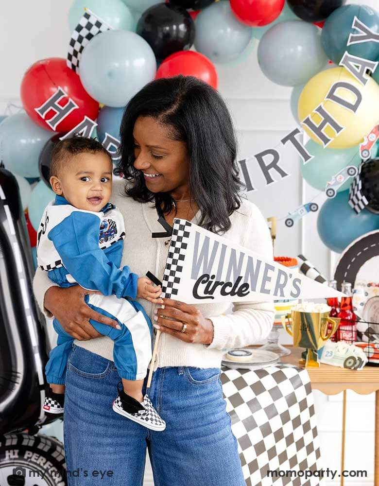 A mom holding his one year old boy in her arm standing in front of a race car themed party decoration set up which includes a colorful balloon garland in red, blue, gray black and yellow adorned with a happy birthday paper banner, on the table there are race car themed partyware including plates, napkins, checkered table runner and race car flags. The mom is holding a felt party pennant written "Winner's Circle" from Momopartys.