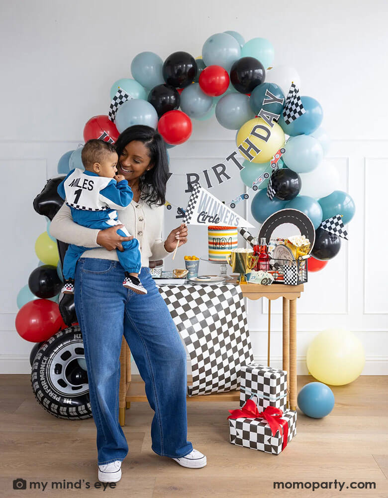 A mom holding his one year old boy in her arm standing in front of a race car themed party decoration set up which includes a colorful balloon garland in red, blue, gray black and yellow adorned with a happy birthday paper banner, on the table there are race car themed partyware including plates, napkins, checkered table runner and race car flags. The mom is holding a felt party pennant written "Winner's Circle" from Momopartys. On the floor there are some birthday gifts wrapped with checkered paper.