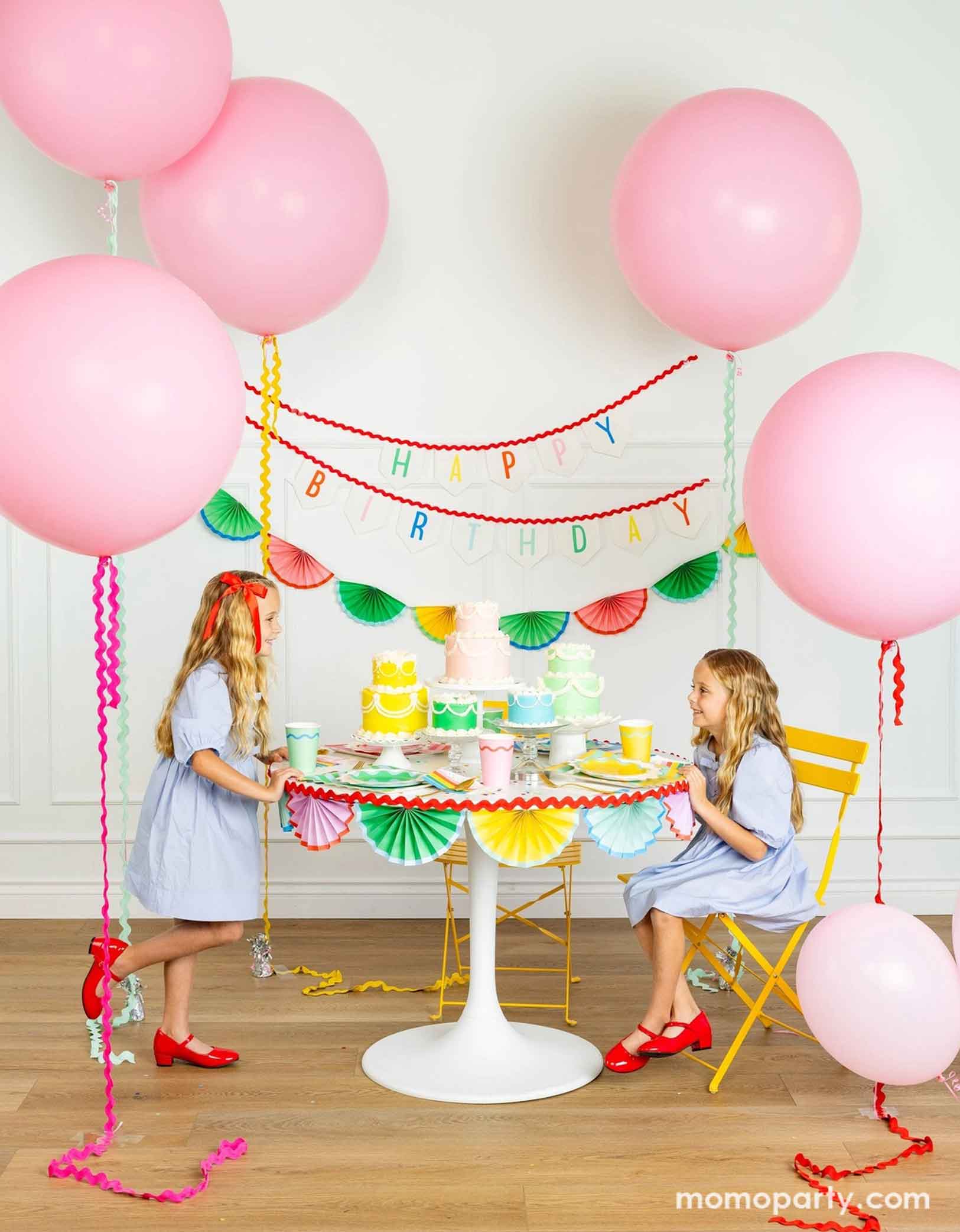 A cheerful and colorful birthday party set up featuring various festive birthday party supplies from Momopartys including rainbow colored tablewares of plates, cups, napkins on the table along with multiple pastel colored birthday cakes. The table is decorated with colorful bunting garland around and in the back of the wall hung a Happy Birthday felt garland and the colorful bunting banner. On the sides are some jumbo pink balloons floating above and two girls in baby blue dresses sitting at the table.
