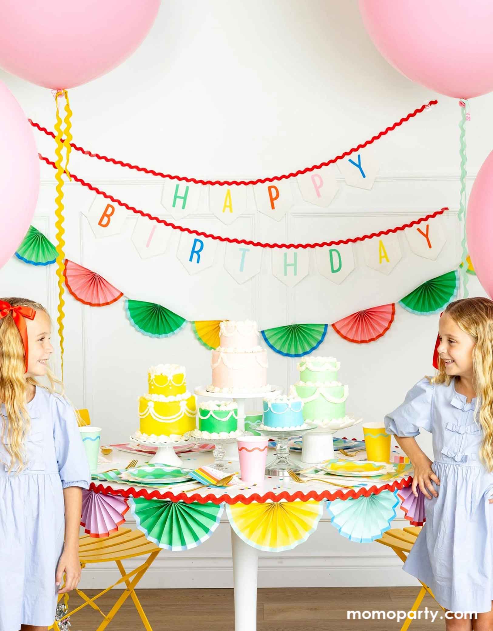 A cheerful and colorful birthday party set up featuring various festive birthday party supplies from Momopartys including rainbow colored tablewares of plates, cups, napkins on the table along with multiple pastel colored birthday cakes. The table is decorated with colorful bunting garland around and in the back of the wall hung a Happy Birthday felt garland and the colorful bunting banner. On the sides are some jumbo pink balloons floating above and two girls in baby blue dresses next to the table.