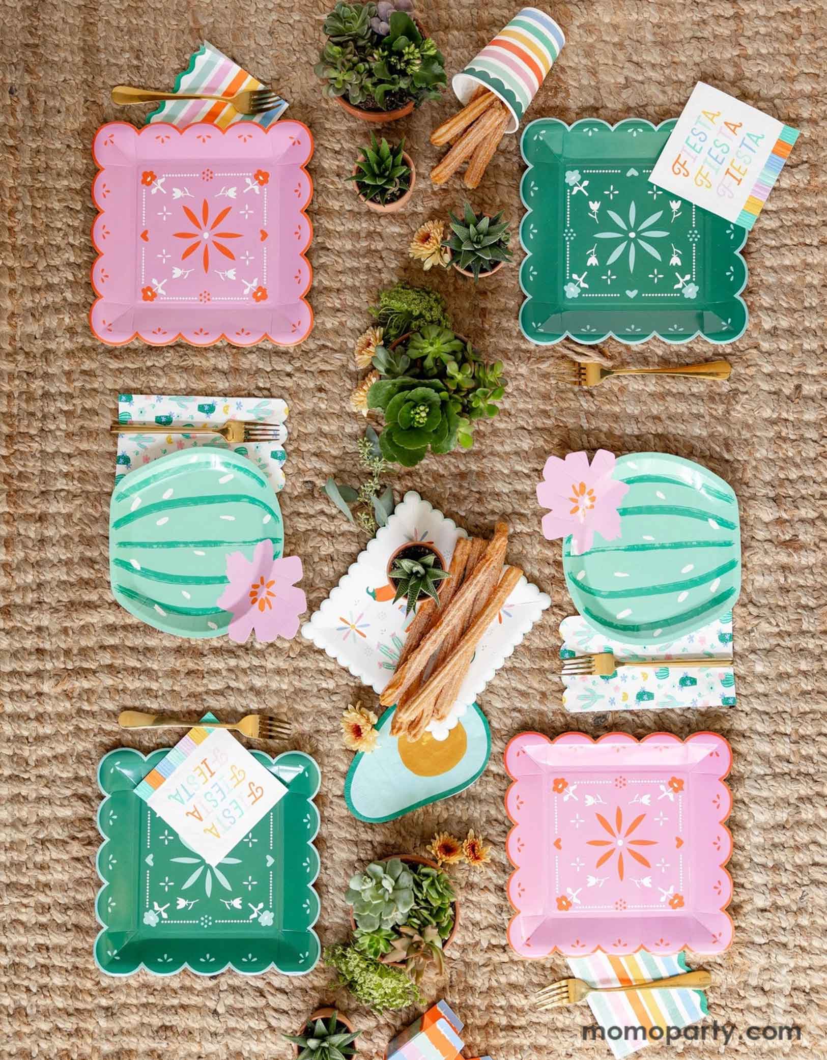A overhead shot of a fun and playful fiesta themed party table featuring Momopartys's fiesta themed party supplies including the Fiesta Papel paper plates in mint and pink, the cactus shaped plates, fiesta fringe napkins, avocado shaped napkins and the fiesta rainbow colored striped party cup filled with churros. On the table there are mini pots of cactus and succulent plants, making this a fun table setting idea for a playful yet modern fiesta themed celebration!