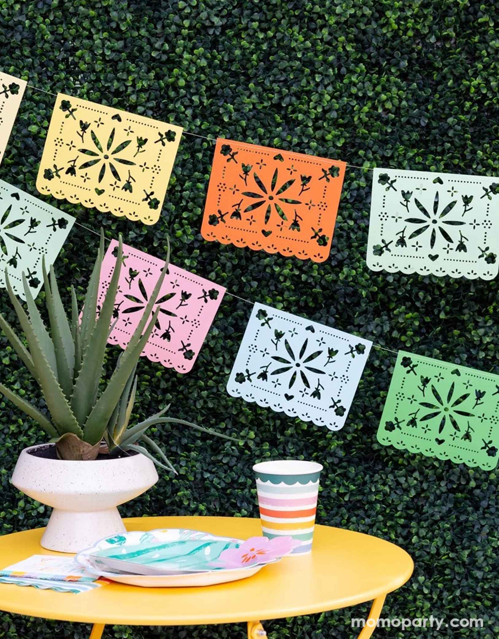 A festive Mexican themed celebration set up featuring Momopartys's Fiesta party supplies including the rainbow colored Fiesta Papel Picado Banner on the faux leaf wall, cactus shaped plate, fiesta fringe napkins and fiesta striped party cups on a bright yellow table. With a cactus/succulent plate as the centerpiece, this makes a simple yet modern look for a fun and playful fiesta celebration!
