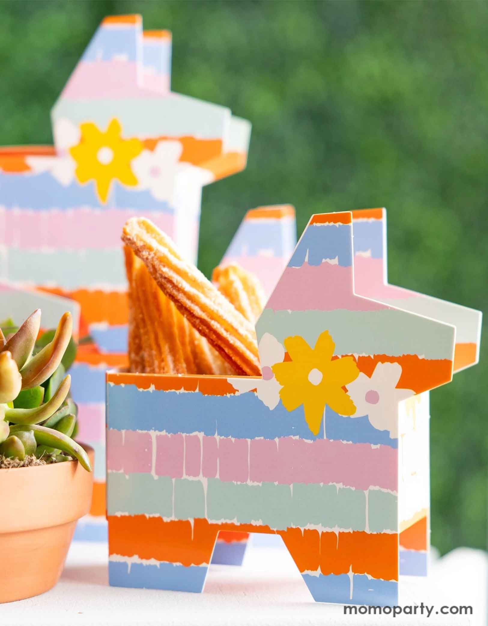 A festive Feista party table featuring Momopartys's fiesta pinata shaped treat boxes in colorful rainbow colors with fringe design. The treat boxes are filled with churros and are set next to a cactus plant, making them a perfect addition to a fun and playful kid's fiesta themed birthday celebration or a Cinco de Mayo party!