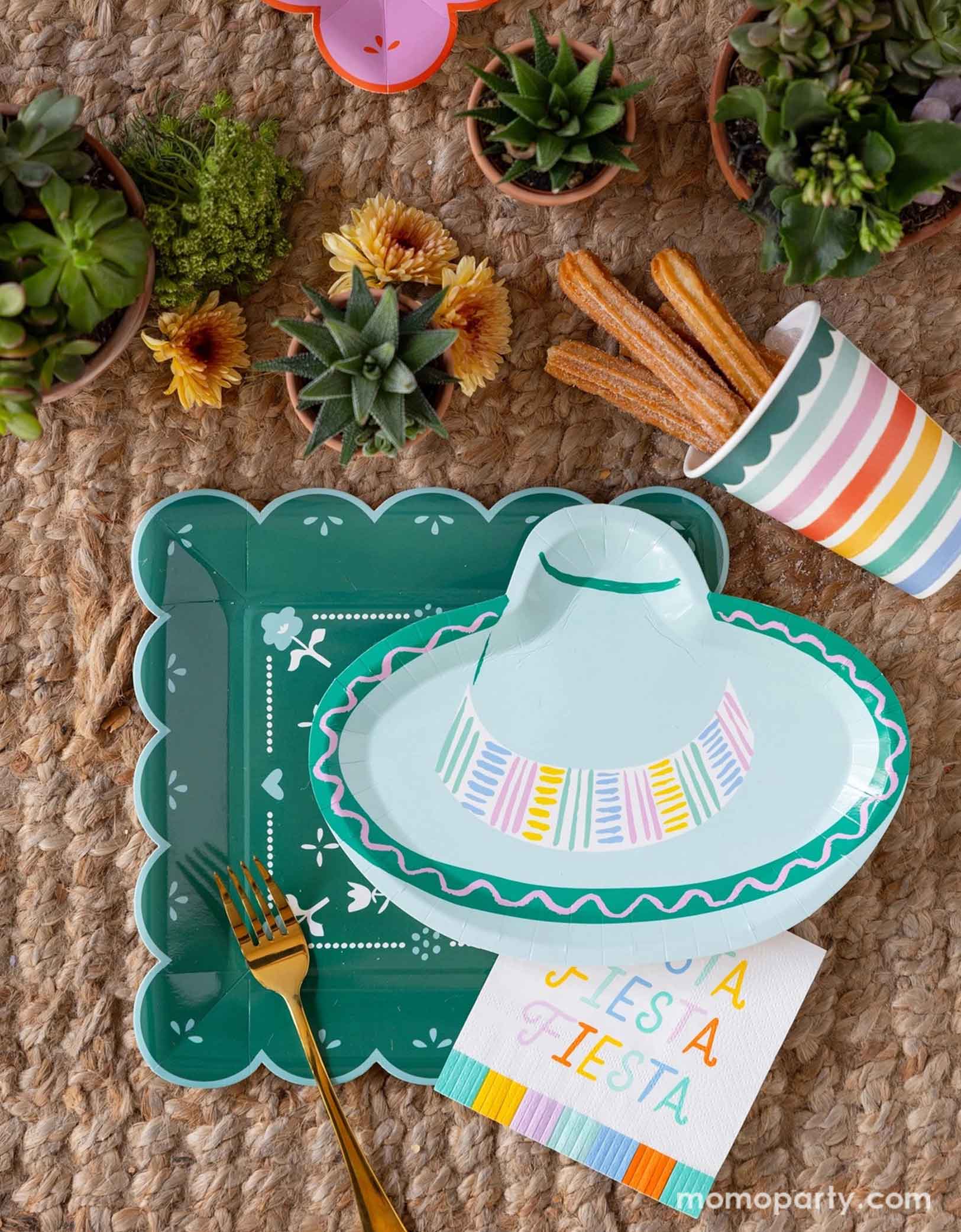 A overhead shot of a fun and playful fiesta themed party tablescape featuring Momopartys's fiesta themed party supplies including the Fiesta Papel paper plate, the sombrero hat shaped plate, fiesta fringe napkins, and the fiesta rainbow colored striped party cup filled with churros. On the table there are mini pots of cactus and succulent plants, making this a fun table setting idea for a playful yet modern fiesta themed celebration - perfect for kid's "first fiesta" themed first birthday party!