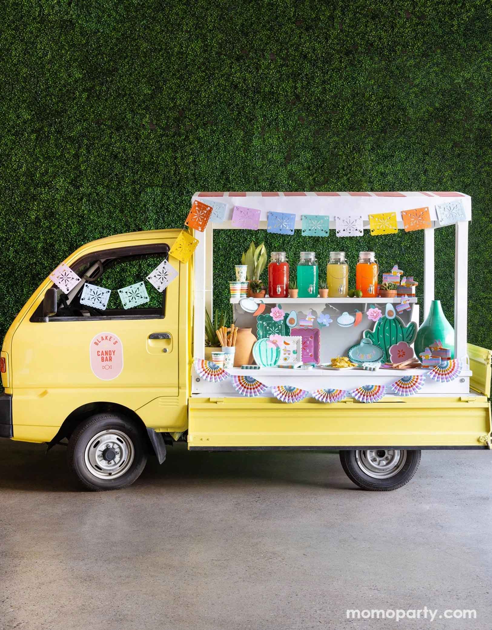 A cheerful yellow Mexican inspired truck is decorated with Momopartys's colorful fiesta party supplies including the Fiesta Papel Picado Banner, bunting banner hung across the truck. In the middle there a various fiesta party tablewares in display including cactus shaped plates, sombrero hat shaped plates, fiesta icon dinner plates, fringe napkins, striped party cups and pinata shaped treat boxes. On the truck there are also colorful drinks in glass pitchers ready to be served for the party guests.