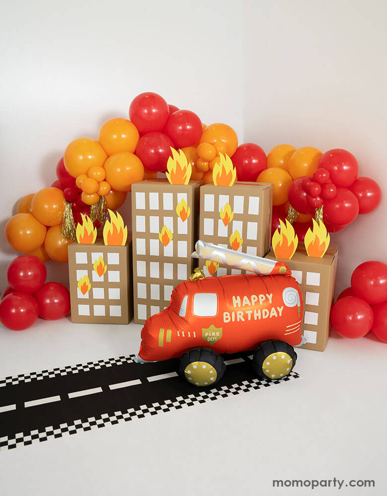 A fire truck themed birthday party balloon decoration featuring Momopartys's fire truck foil balloon and balloon garland in red and orange. Under the balloon garland there are playhouse buildings made out of packaging boxes with fire flame paper cutouts, making this a fun and playful decoration for kid's fire truck themed birthday party or classroom decoration!