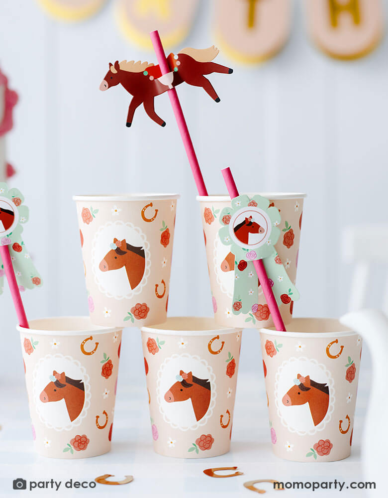 A stack of Momopartys's 6.7 oz floral pony party cups featuring adorable pony, floral and horseshoe design in blush color. There are also the floral pony paper straws in pink with paper pennants. On the table you can see some gold horseshoe shaped confetti, making this a perfect inspiration for kid's pony or horse themed birthday party set up.