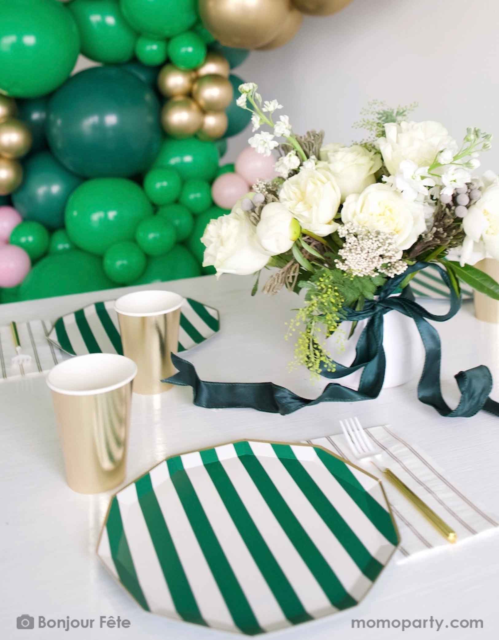 An elegant football themed party table featuring Momopartys's emerald green and cabana stripe and gold tableware including plates, party cups, napkins and gold utensils. With an elegant flower bouquet in white and green with a dark green satin ribbon bow on the vase, along with a balloon wall that consists of green, pink and gold balloons, this is a party inspiration for a girly football, Super Bowl, or a sports themed celebration.