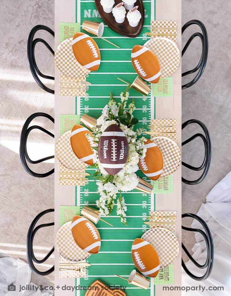 A overhead shot of a long dining table featuring Momopartys's football themed party supplies, including the die-cut football shaped plate, gold and white checkered dinner plate with green and coordinating napkins and gold party cups. In the center of the table there's a football field table runner and a beautiful flower bouquet, all makes great inspirations for football themed birthday celebrations or a Super Bowl, tailgate party.