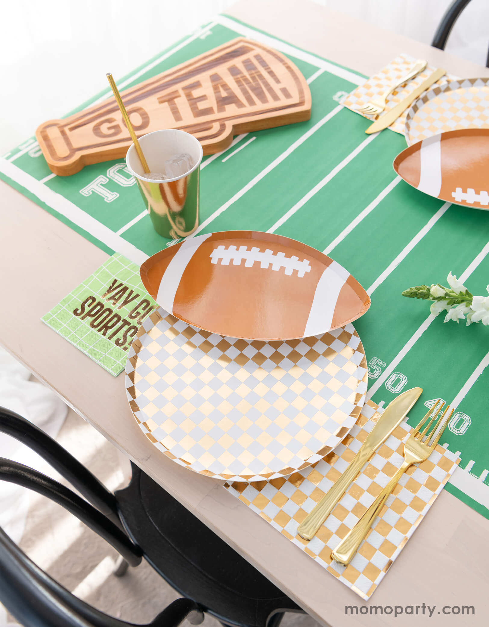 A modern football party table filled with Momopartys's football themed party tableware by Daydream Society including the die-cut football shaped plate, gold and white checkered dinner plate with green napkins says YAY GO SPORTS and gold party cups. In the center of the table there's a football field table runner and a megaphone shaped snack board says GO TEAM! Making this a great inspirations for football themed celebrations, a Super Bowl watching party or a Sunday football viewing party.