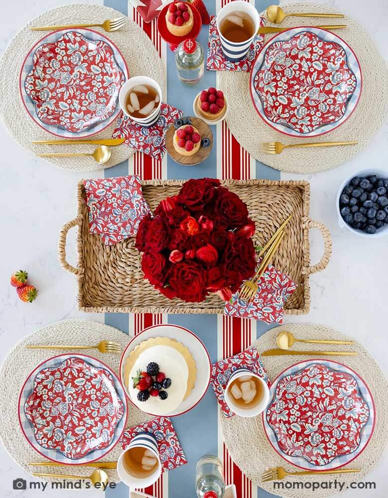 An elegant patriotic American themed party table features Momopartys's Hamptons Party Collection by My Mind's Eye including 10" Chambray Gingham Paper Plates, 10" Hampton Floral Wave Plates and napkins on white round placemats. In the middle of the table there are blue and red cakes, treats and drinks, along with a red floral bouquet as the centerpiece in the center on the Hampton striped paper runner, making this a perfect tablescape inspo for a 4th of July party or Memorial Day celebration.