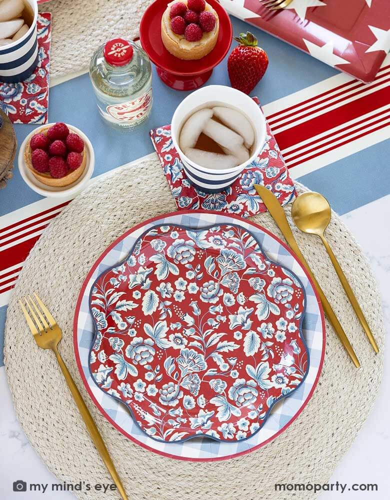 An elegant Fourth of July party table features Momopartys's Hamptons Collection by My Mind's Eye including 10" Chambray Gingham Paper Plate, Hampton Floral Wave Plate and napkin on a round white placemat. In the middle of the table there are blue and red treats and drinks on the Hampton striped paper runner, making this a perfect party inspo for a 4th of July party or Memorial Day celebration.