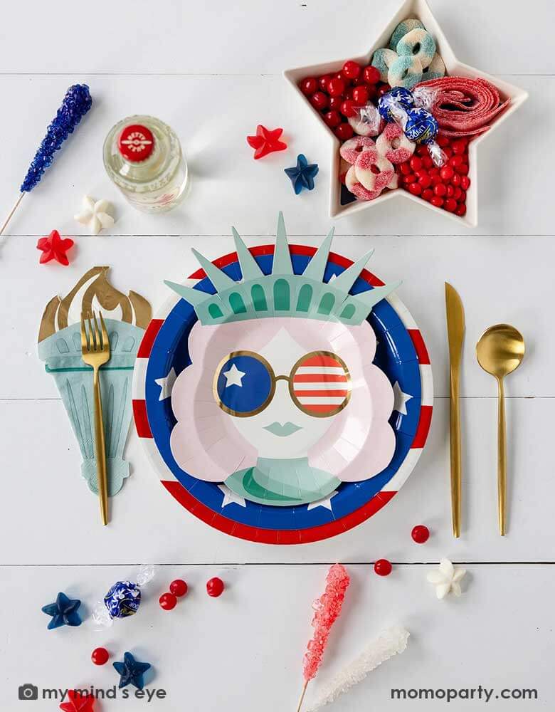 A modern and fun placeset featuring Momopartys's Lady Liberty Party Collection by My Mind's Eye including the 10" red striped round plate, 9" blue star round plate, lady liberty shaped plate, torch shaped dinner napkins and. Around them are various candies and treats in patriotic themed in red white and blue. A perfect party inspo for a modern Fourth of July celebration with friends and family.